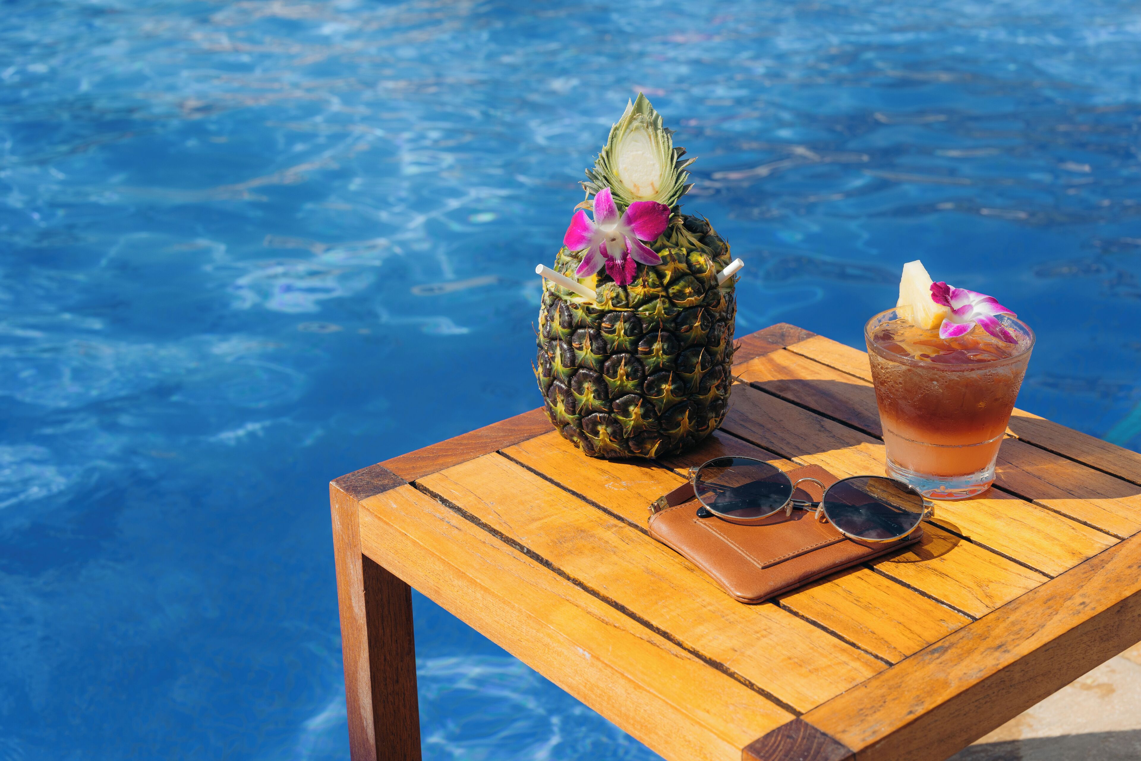 Une table en bois près d’une piscine, avec un ananas, une boisson, des lunettes de soleil et un portefeuille.