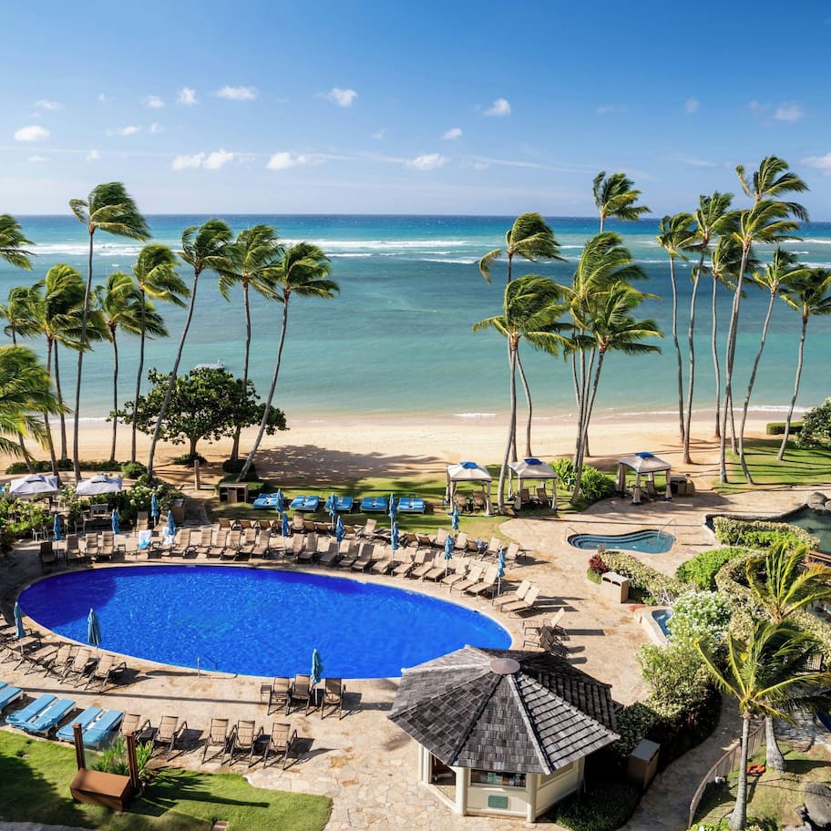 A resort with a swimming pool, beach view, palm trees, and lounge chairs.