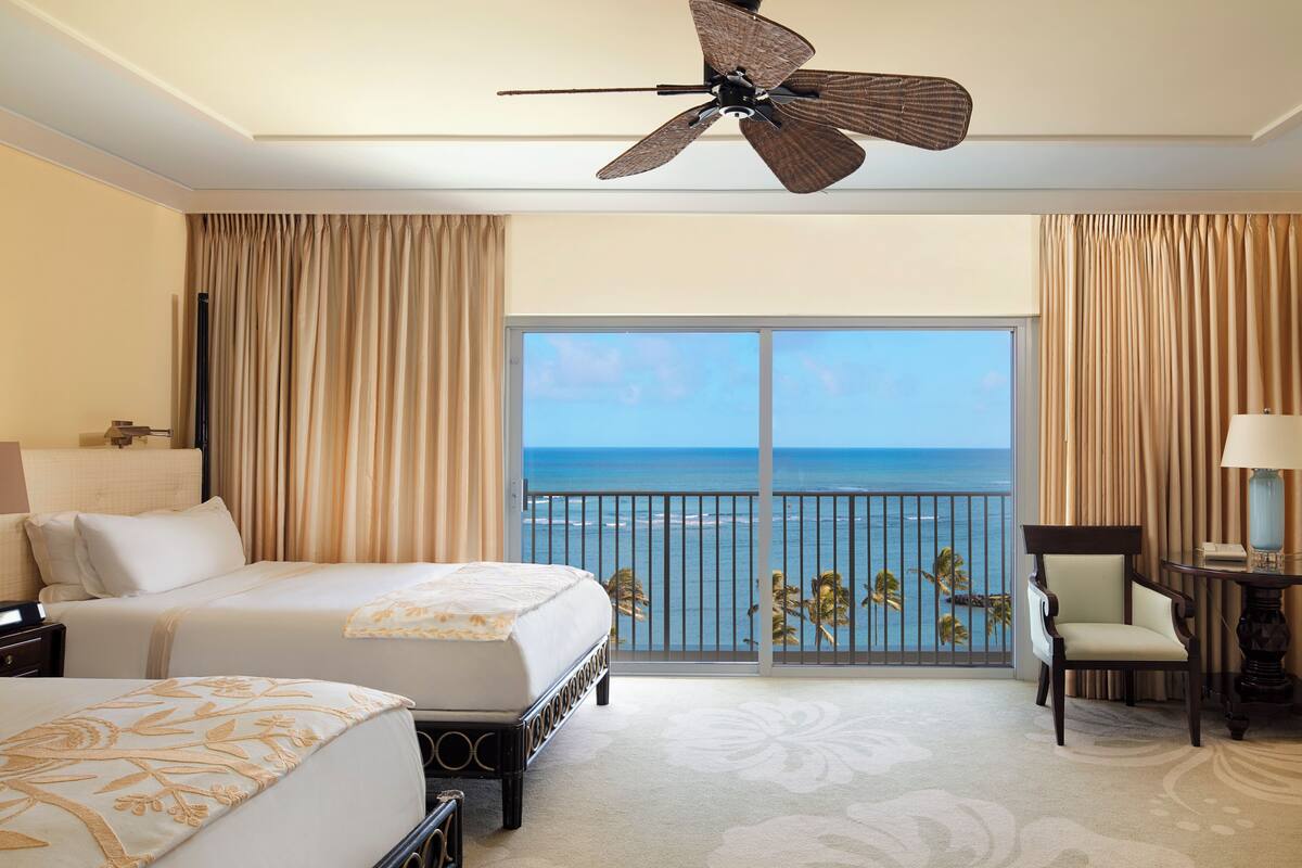 A hotel room with a bed, a chair, a desk with a lamp, and a view of the ocean.