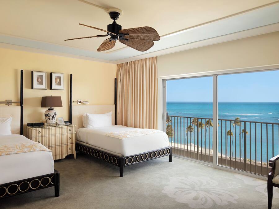 A hotel room with two beds, a ceiling fan, a large window with a view of the ocean, and a balcony with a railing.