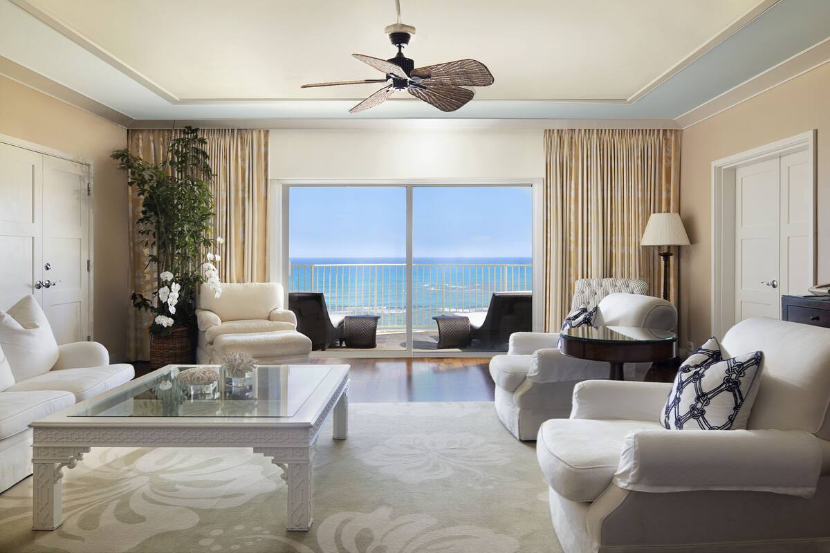 A spacious living room with a large window overlooking the sea, furnished with white sofas, armchairs, and a coffee table.