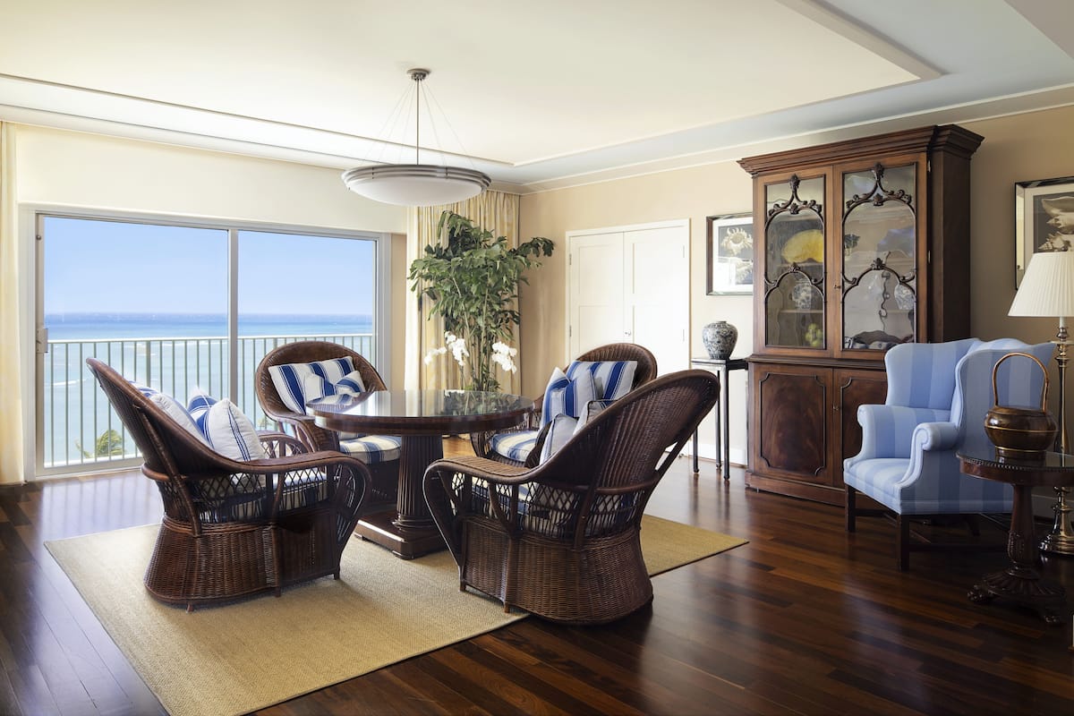 A spacious living room with a large window offering a view of the ocean, wicker furniture, a wooden cabinet, and a blue armchair.