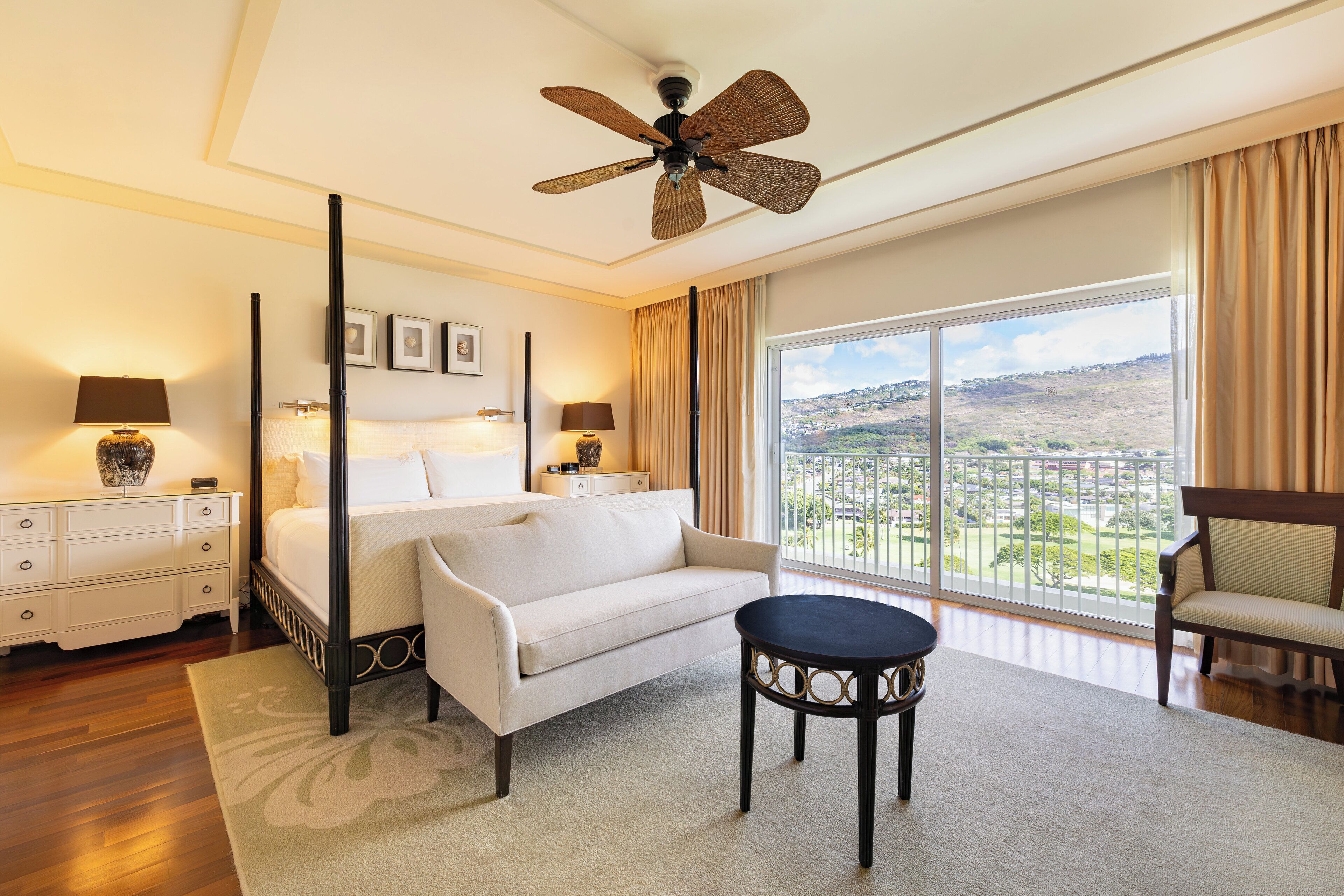 A hotel room with a bed, a sofa, a small table, a ceiling fan, a nightstand, a lamp, and a large window with a view of the landscape.