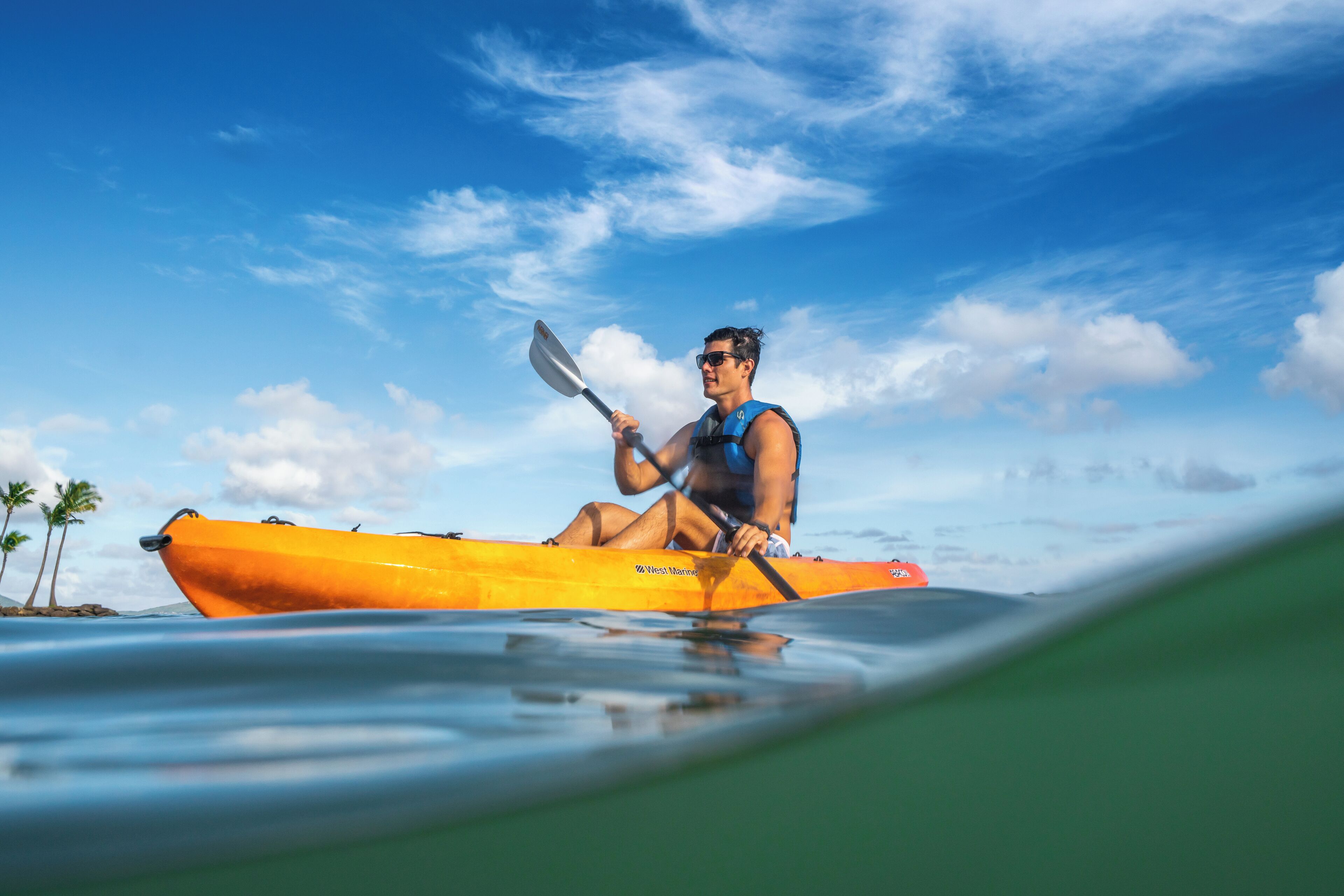 A person in a yellow kayak on calm water with palm trees in the background.