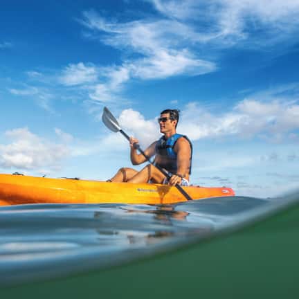 A person in a yellow kayak on calm water with palm trees in the background.