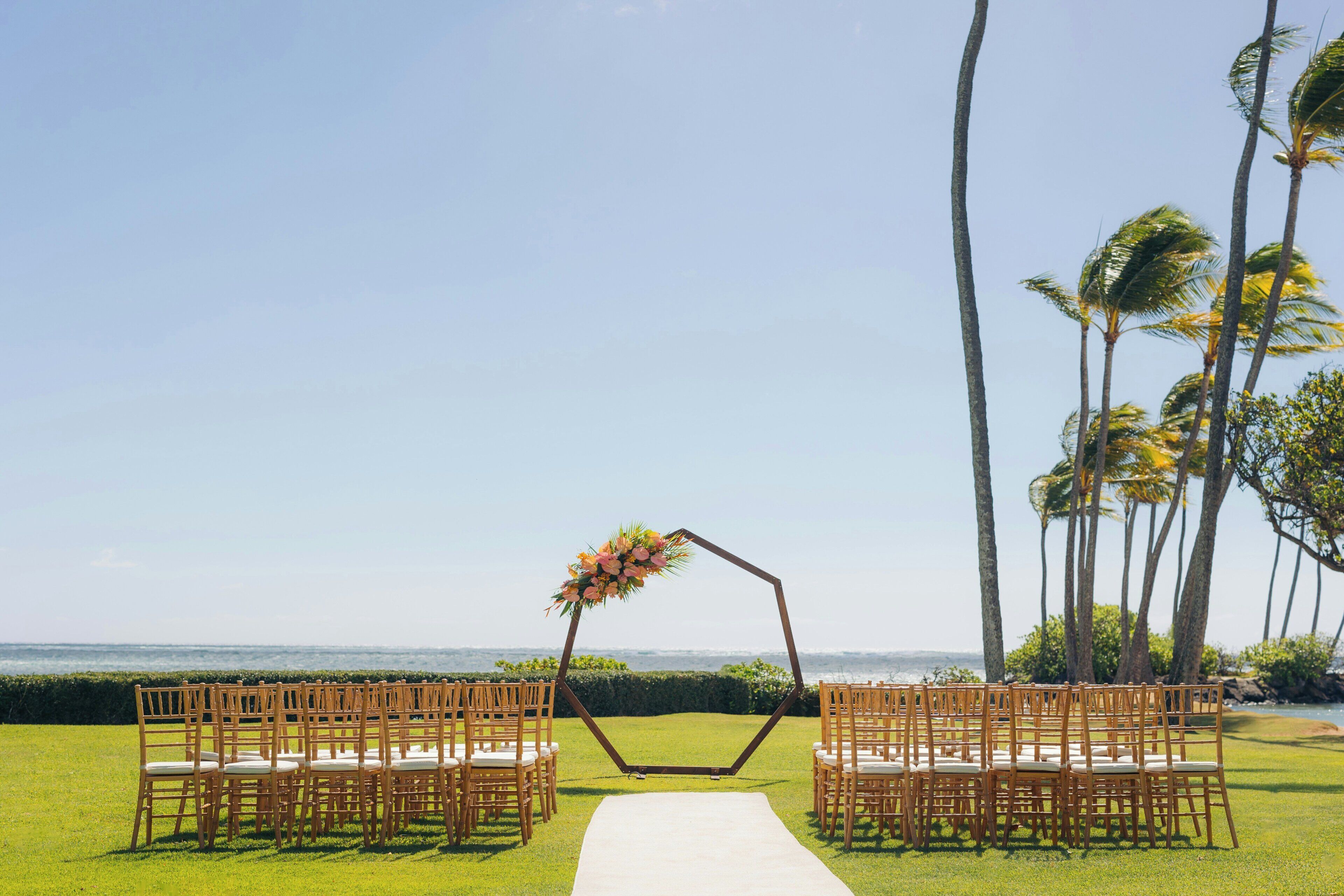 Une cérémonie de mariage avec une arche hexagonale, une allée et des chaises disposées sur une pelouse, le tout offrant une vue sur l’océan.