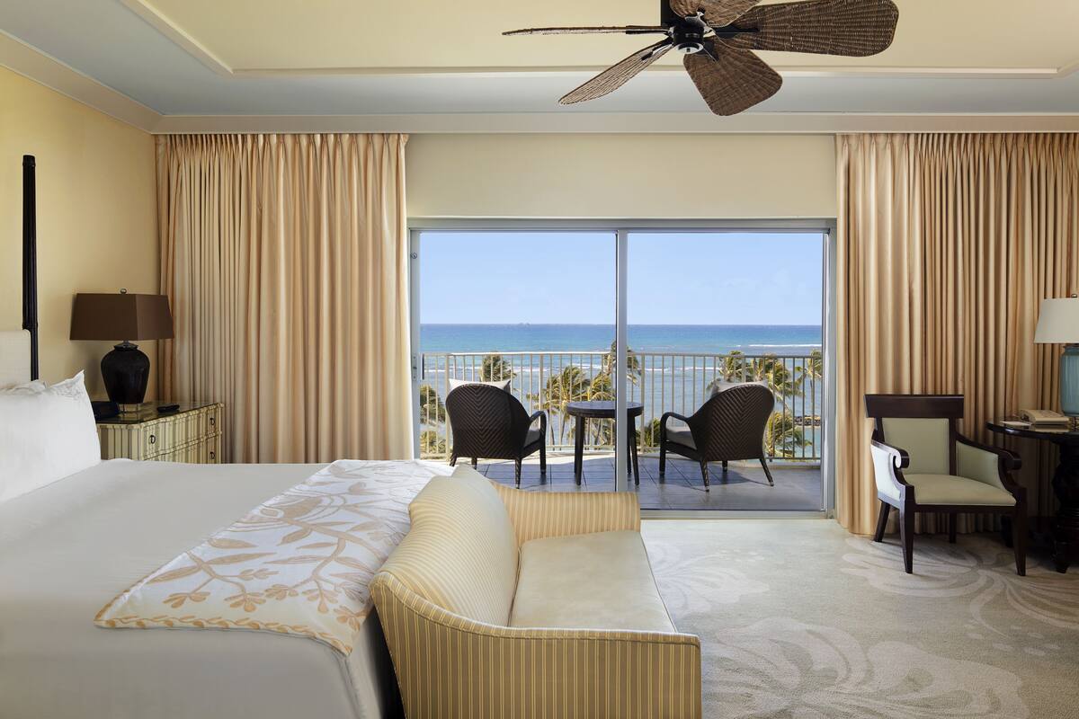 A hotel room with a large bed, a ceiling fan, a balcony with chairs, and a view of the ocean.