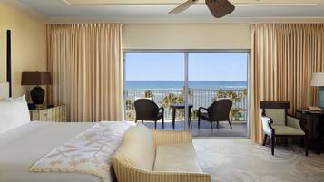 A hotel room with a large bed, a ceiling fan, a balcony with chairs, and a view of the ocean.