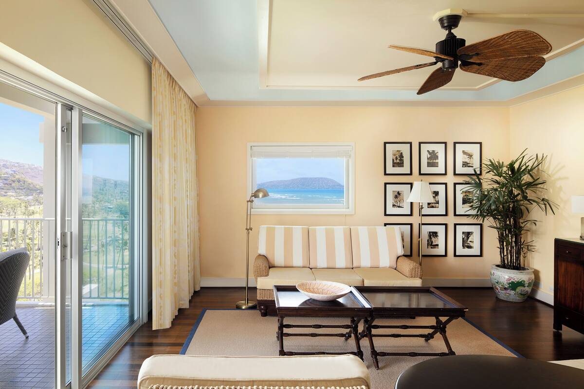 A living room with a sofa, a coffee table, a chair, a large window with a view, a ceiling fan, and a potted plant.