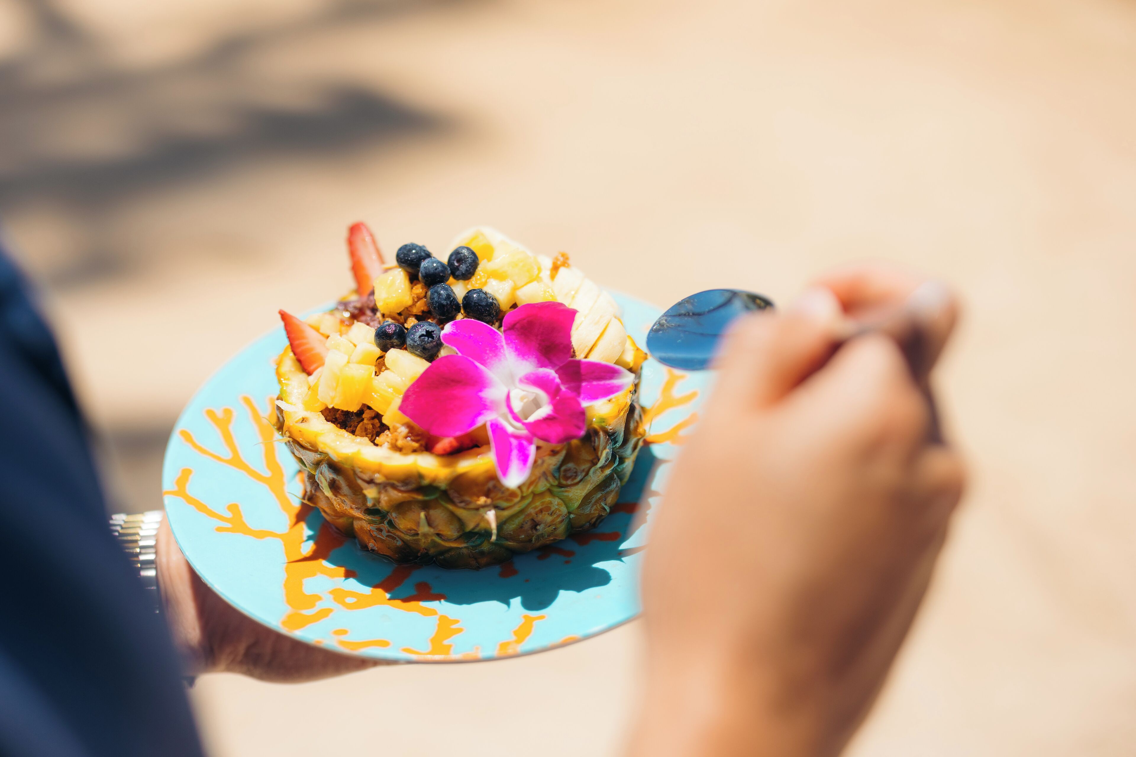 A person holding a plate with a pineapple dessert topped with fruit and a flower.