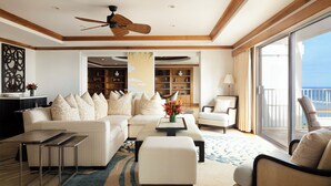 A spacious living room with a sofa, ottoman, coffee table, and a ceiling fan. There is a large window with a view of the ocean.