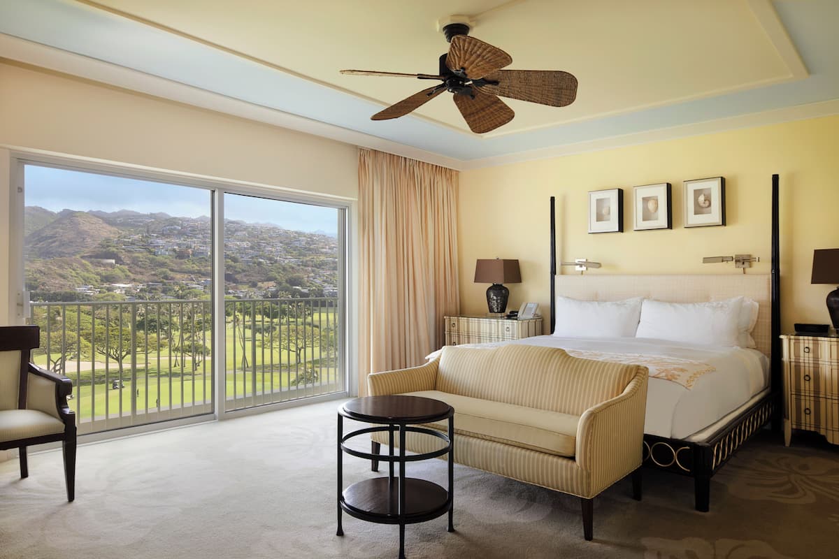 A hotel room with a large balcony, a bed, a chair, a small table, and a ceiling fan.