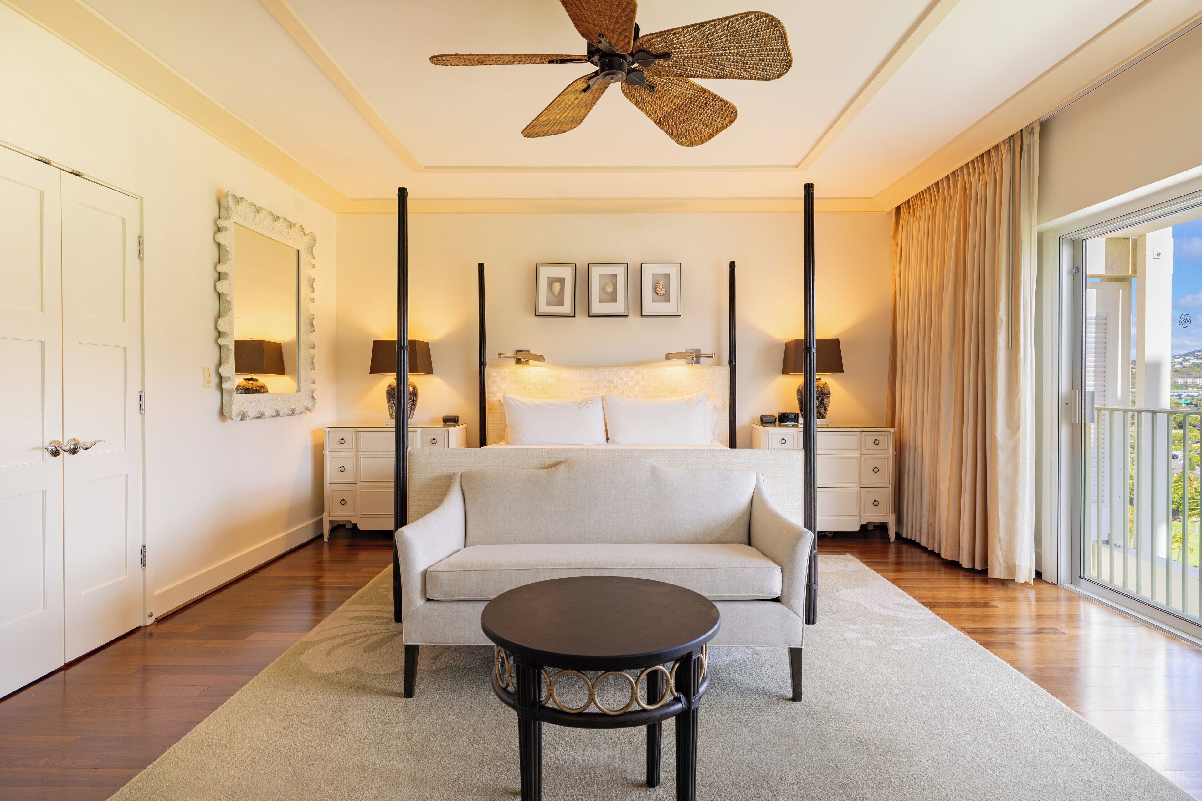 A bedroom with a four-poster bed, a ceiling fan, bedside lamps, a round table, and a view through the window.