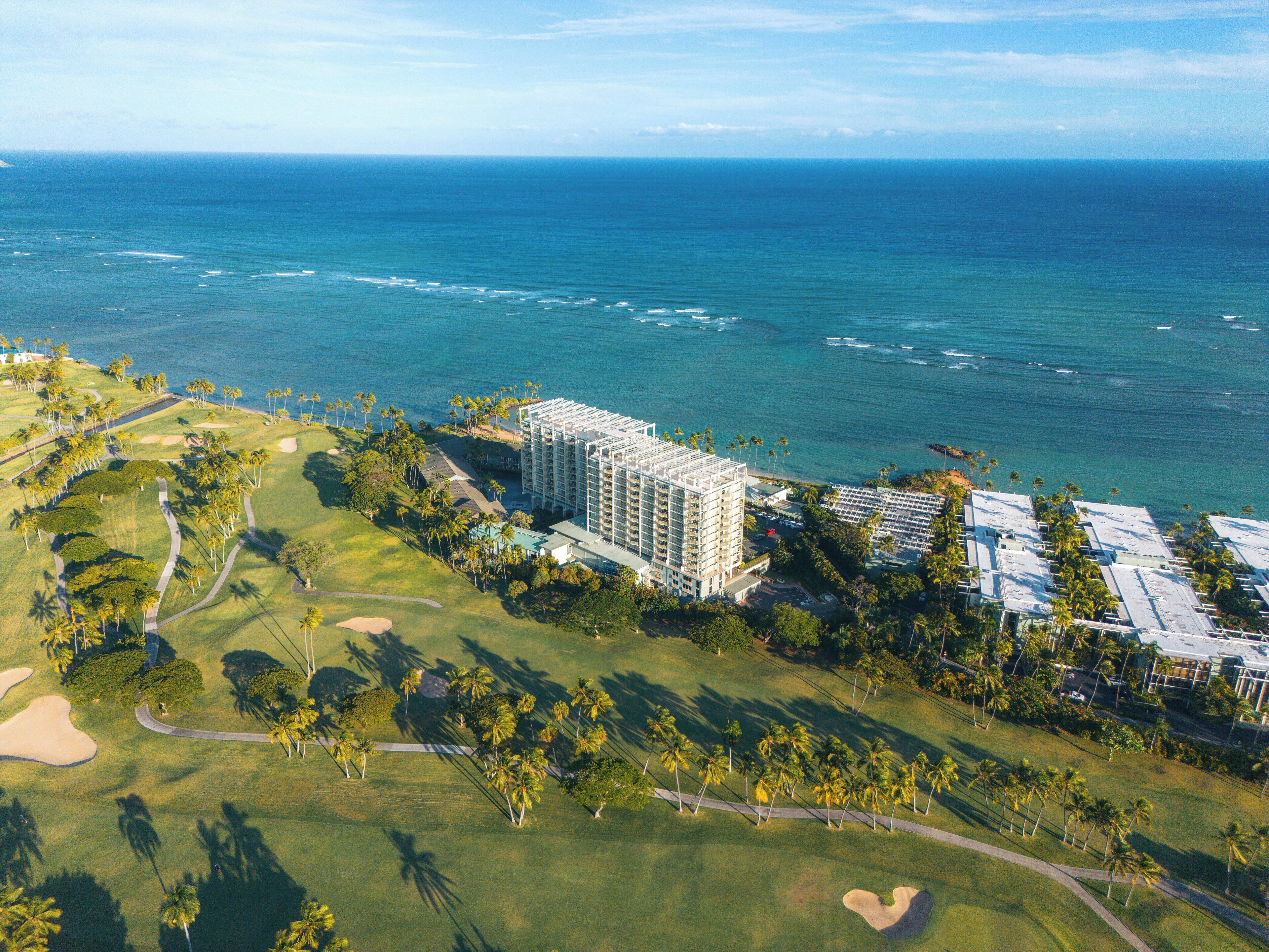 An aerial view of a coastal resort with a golf course, a large hotel building, and a clear view of the ocean.