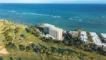 An aerial view of a coastal resort with a golf course, a large hotel building, and a clear view of the ocean.