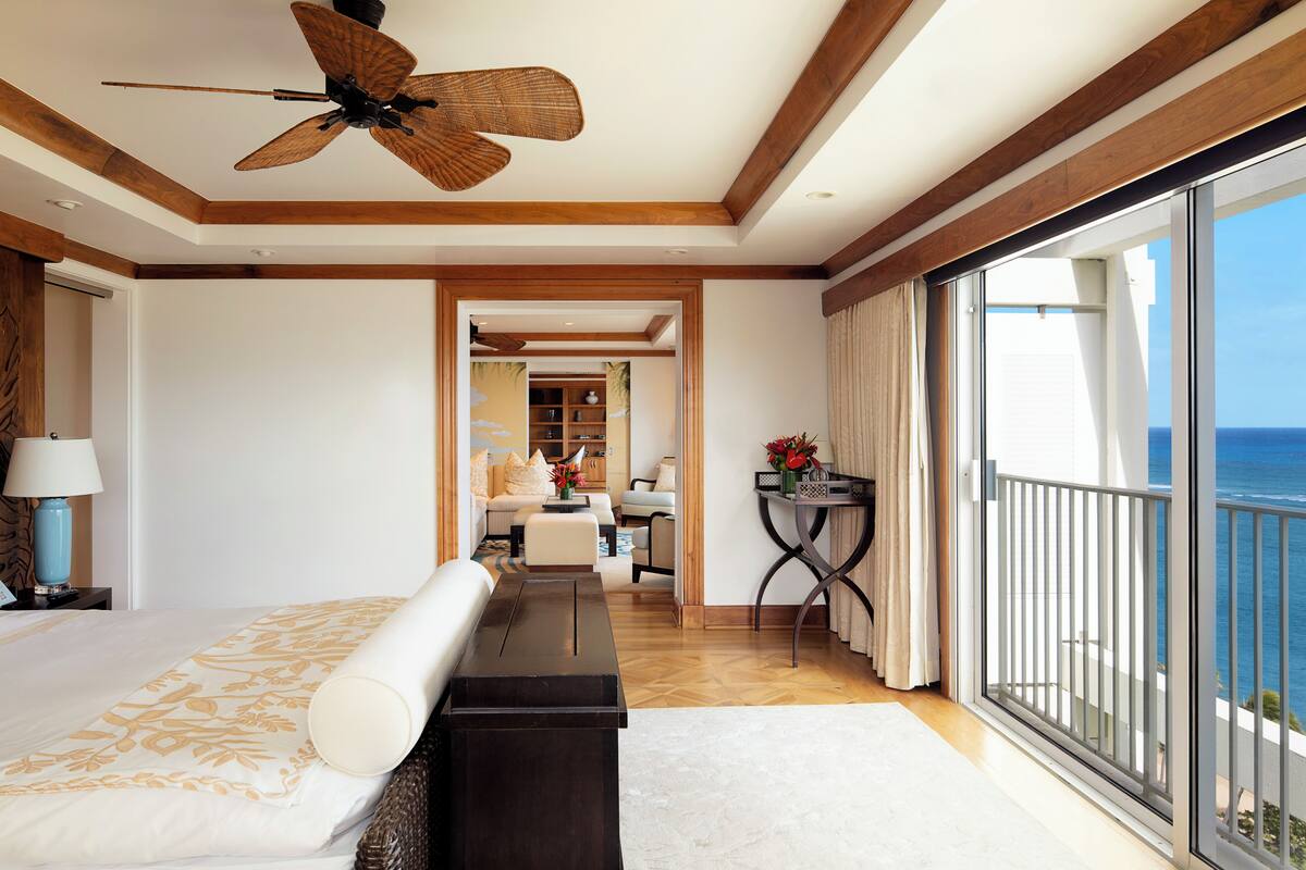 A hotel room with a large bed, a desk, a balcony with a view of the ocean, and a ceiling fan.