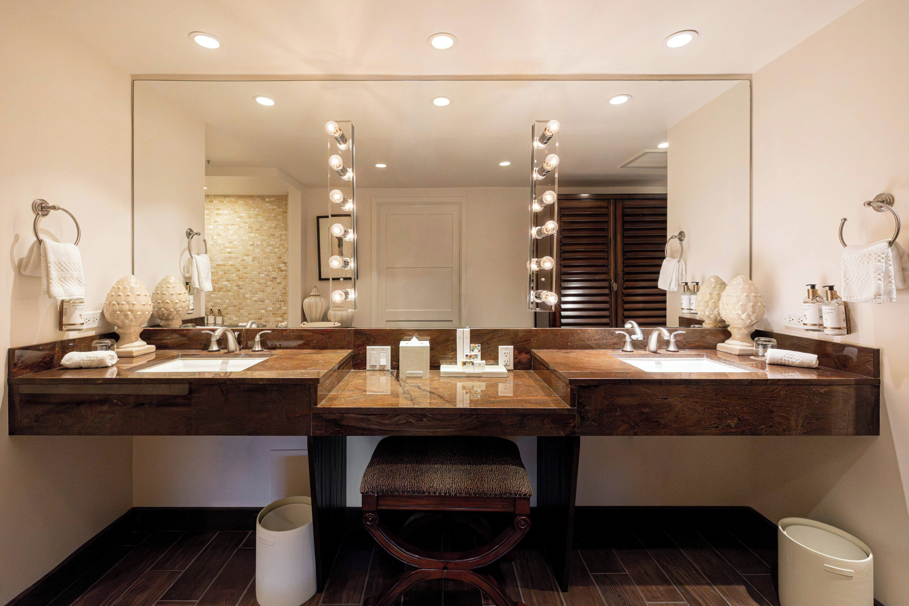 A bathroom with two sinks, a large mirror with lights, a bench, and a trash can.
