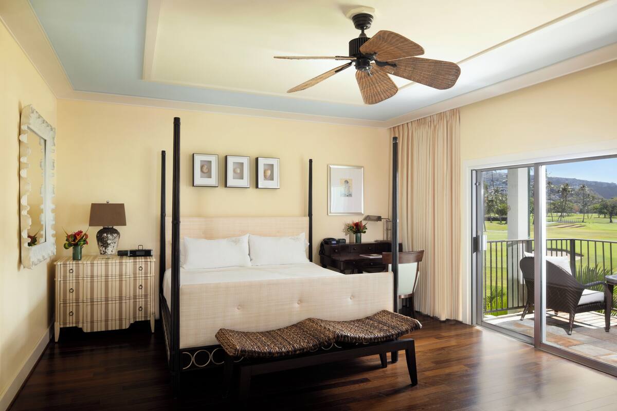 A four-poster bed with a canopy, a bedside table with a lamp, a bench, and a view of a golf course through a sliding glass door.