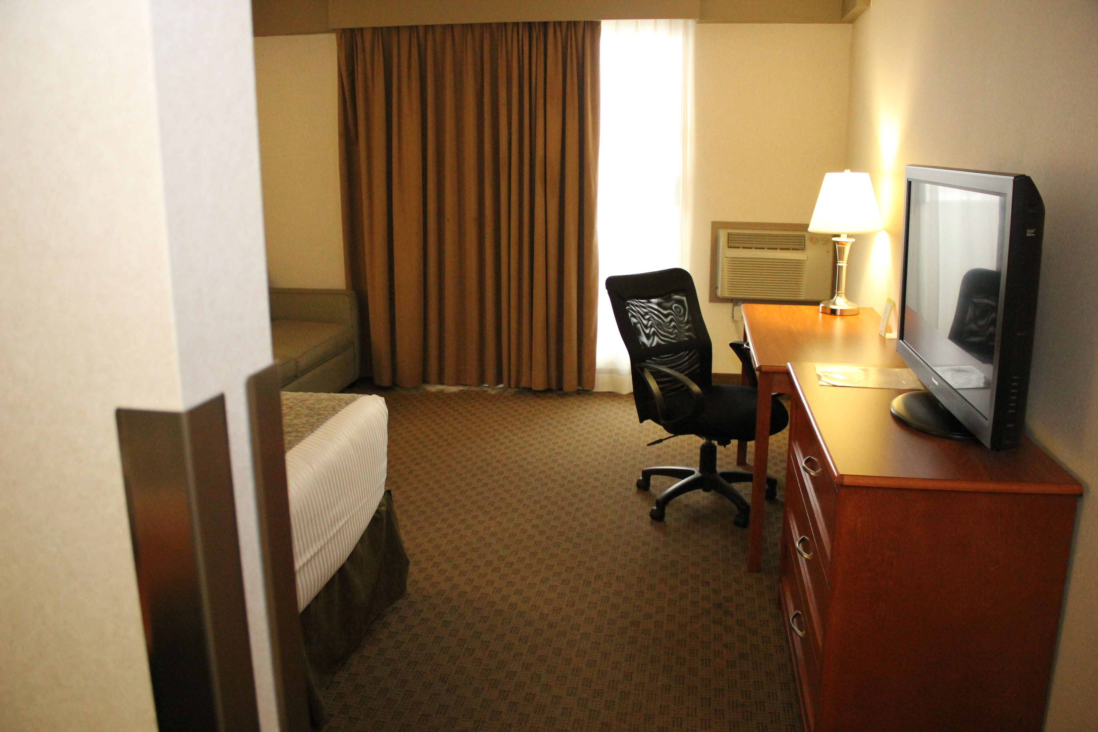 A hotel room with a bed, a desk with a computer monitor, a chair, and a lamp.
