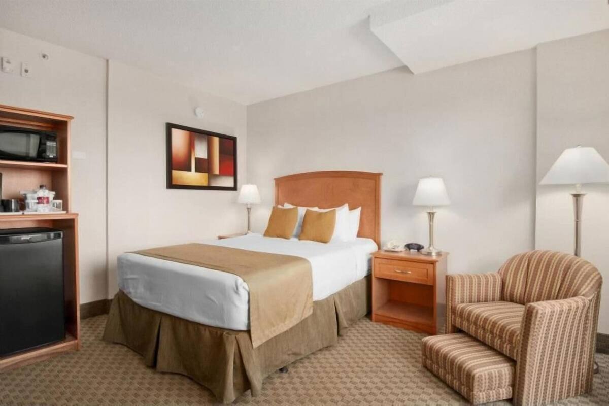 A hotel room with a bed, bedside table, lamp, chair, and a kitchenette area.