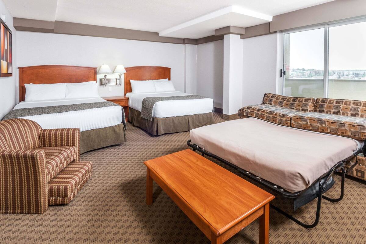 A hotel room with two beds, a sofa, a small table, and a large window offering a city view.