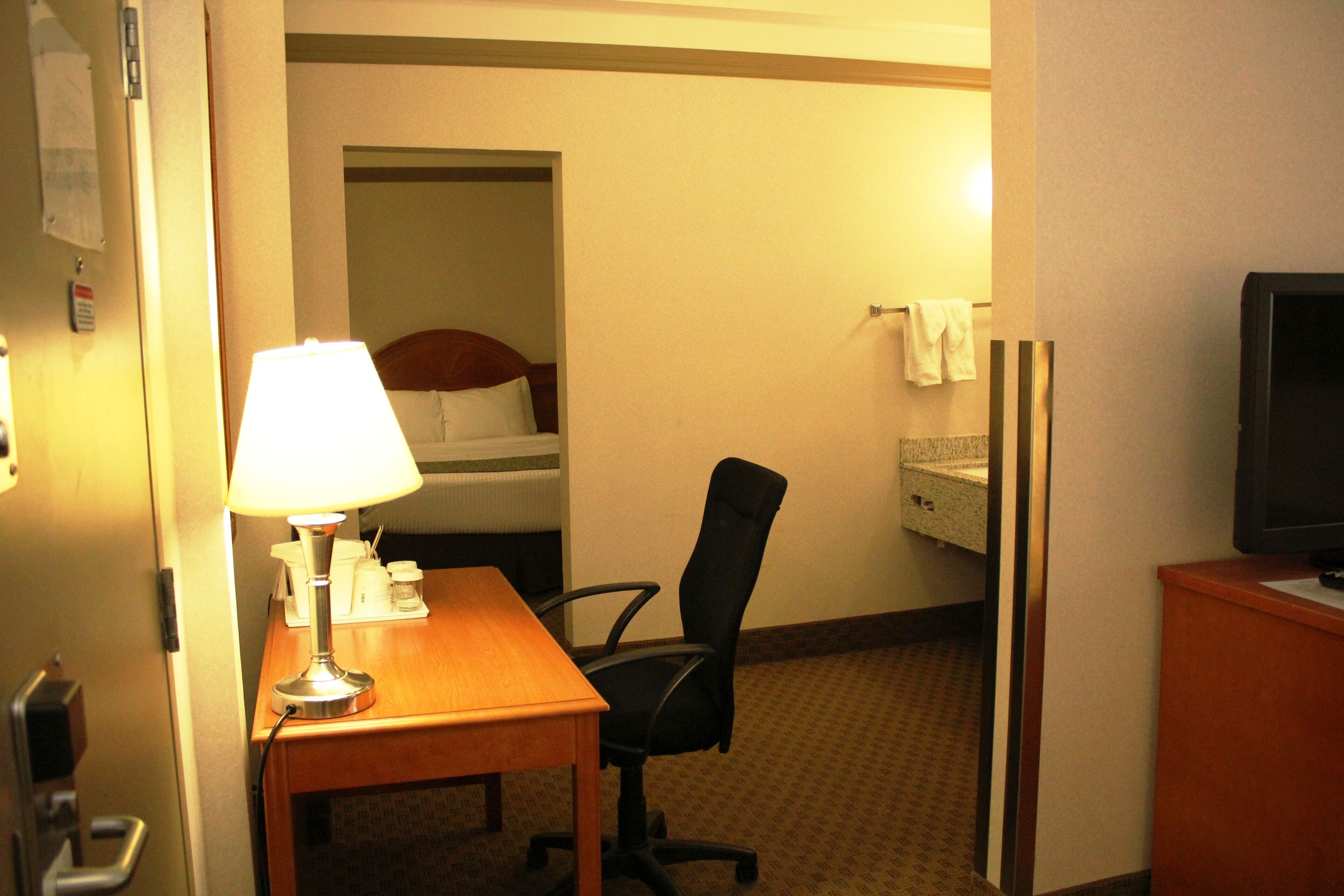 Habitación de hotel con un escritorio de madera, una silla de oficina negra, una cama con ropa blanca, un televisor sobre un mueble de madera y un espejo en la pared.
