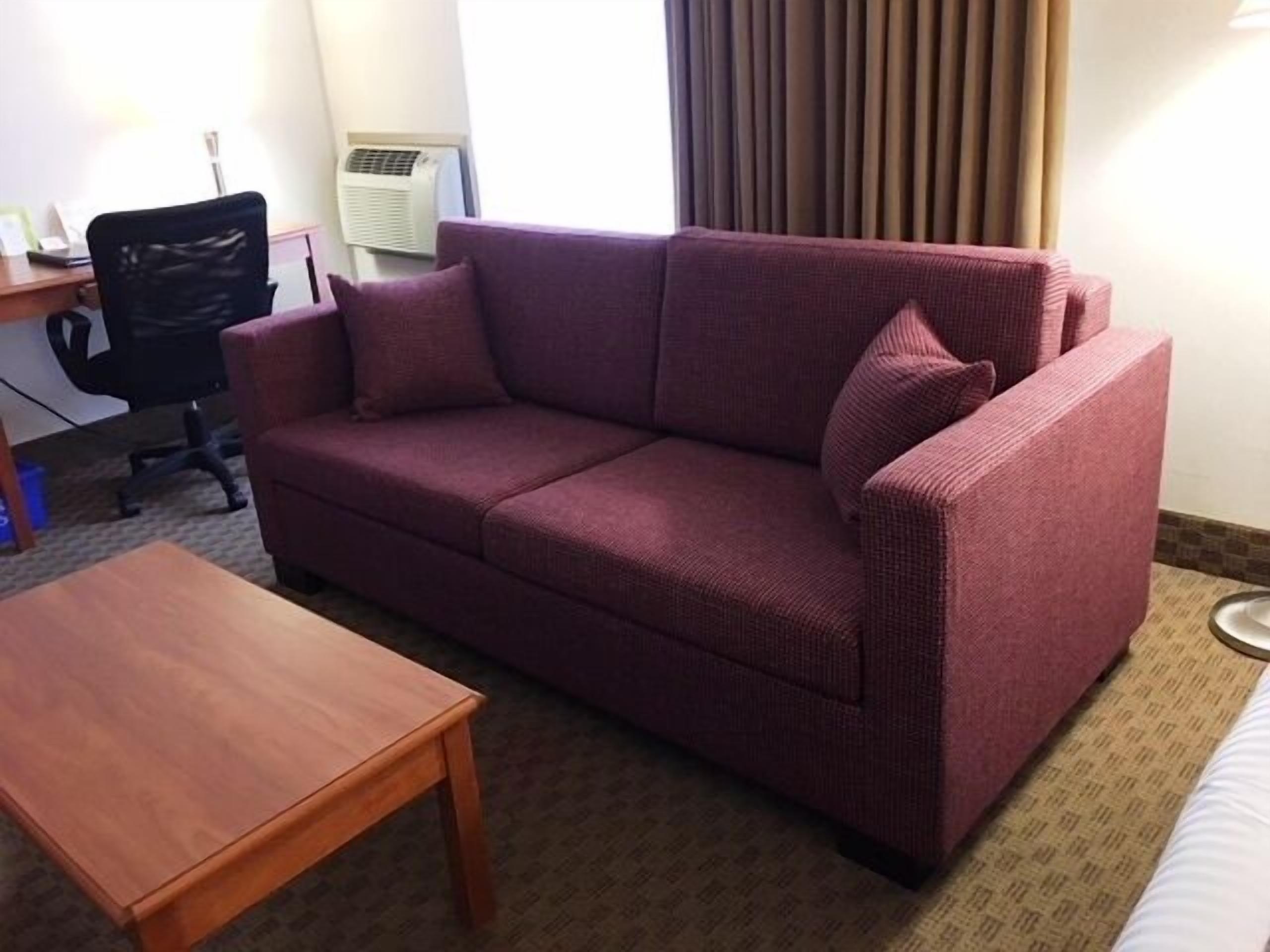 A room with a purple sofa, a wooden coffee table, a black office chair, and a window with curtains.