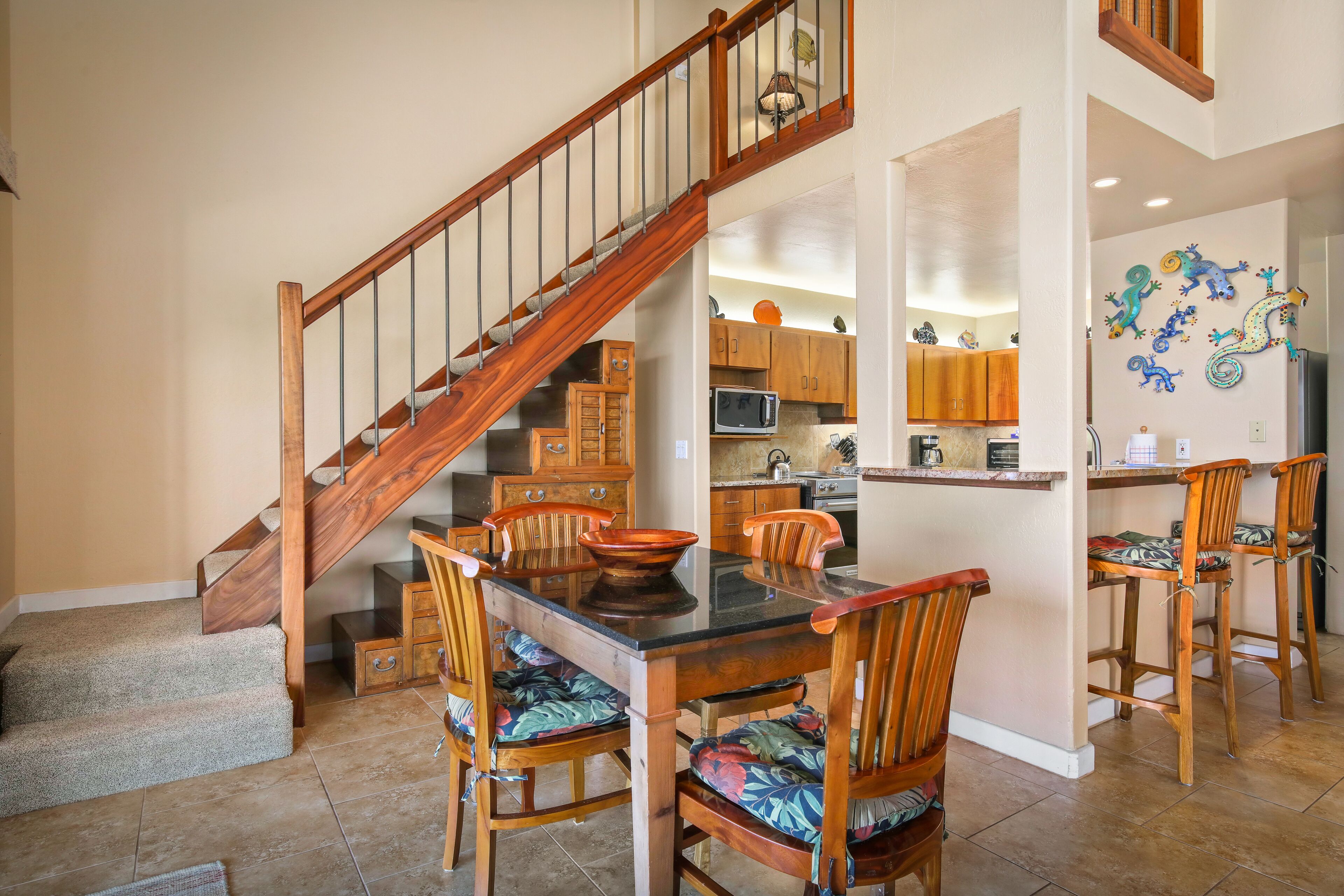 A dining area with a wooden staircase, a kitchen in the background, and a wall with colorful wall art.