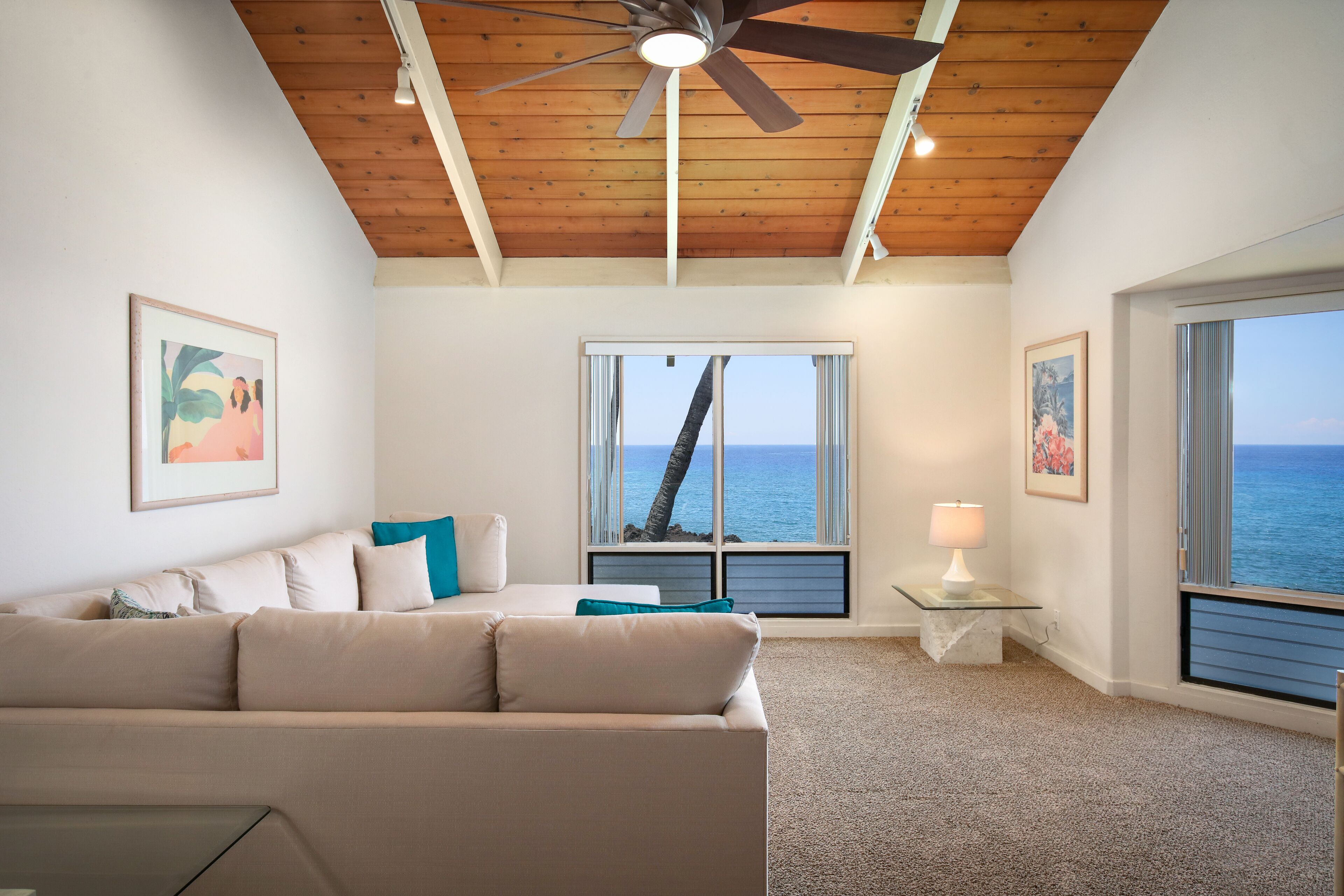 A living room with a beige sofa, a ceiling fan, and large windows with ocean views.