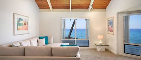 Un salón con un sofá beige, un ventilador de techo y grandes ventanas con vistas al océano.