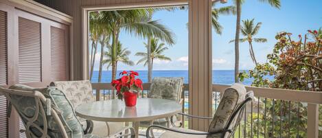 A covered outdoor seating area with a round table and chairs, overlooking a beach with palm trees and the ocean.
