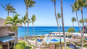 A pool area with lounge chairs and umbrellas, surrounded by palm trees and overlooking the ocean.