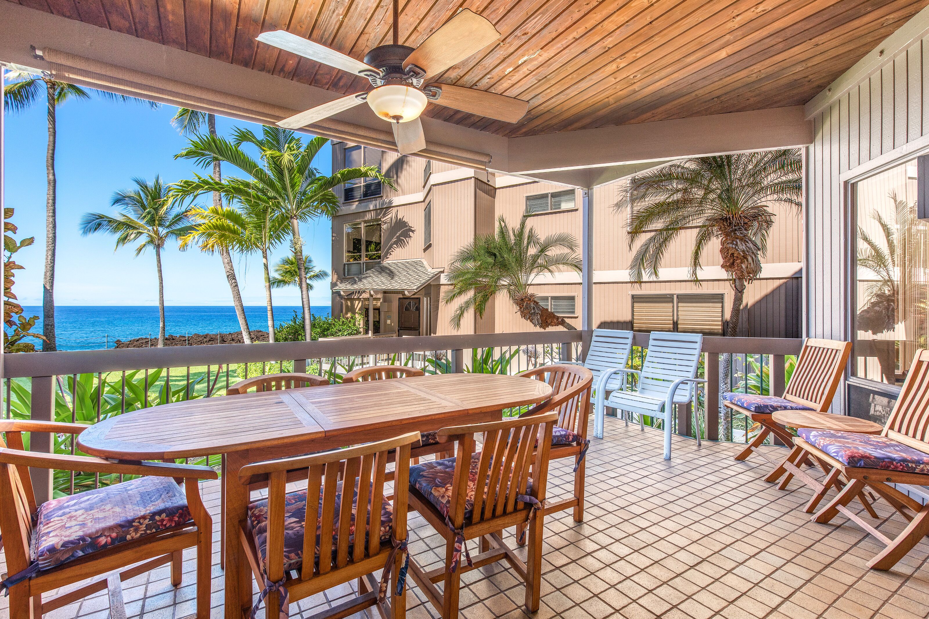 A covered outdoor area with a wooden table and chairs, a ceiling fan, and a view of the ocean.