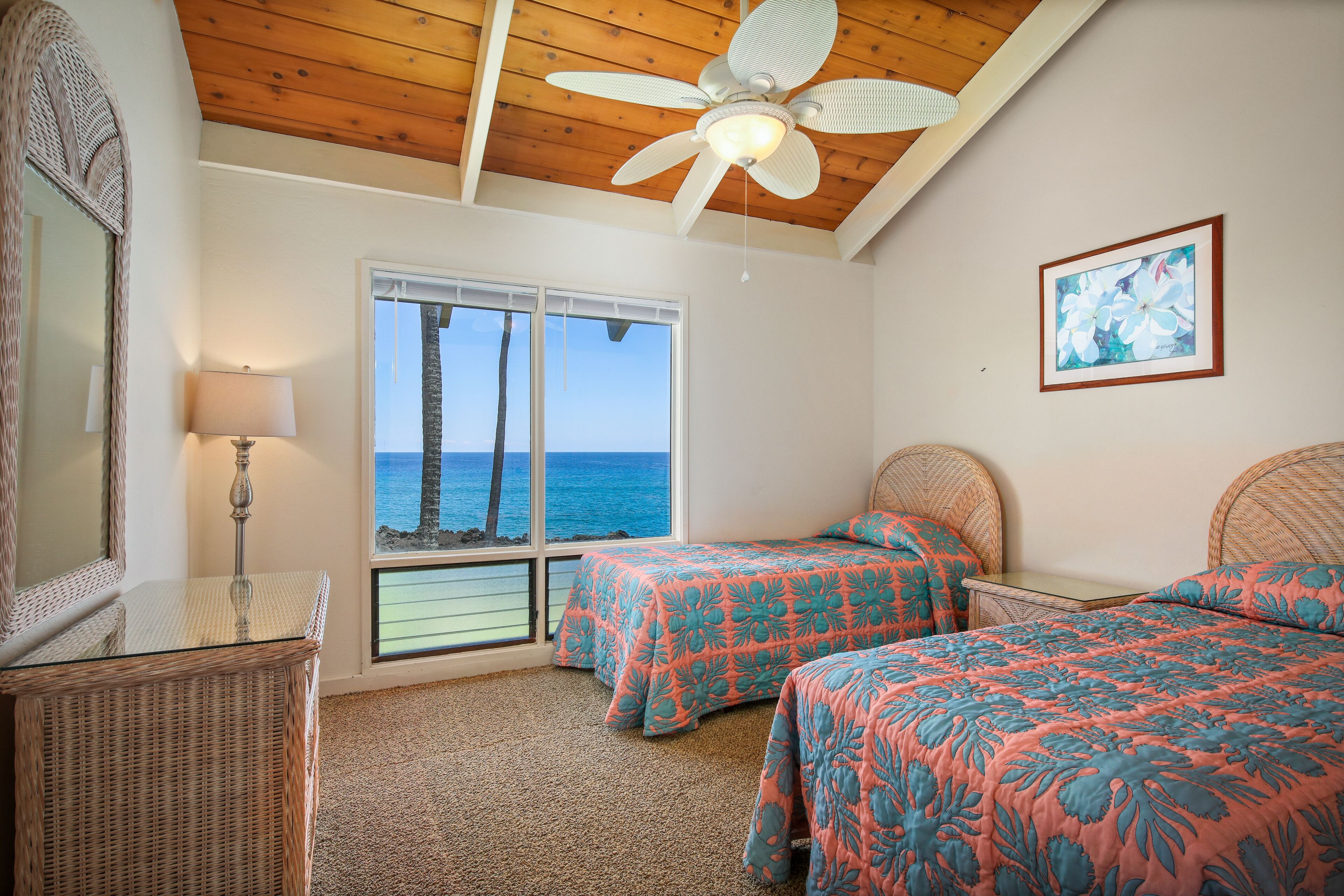 A bedroom with two beds, a ceiling fan, a large window with a view of the ocean, and a framed picture on the wall.