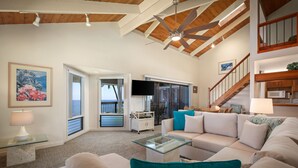 A spacious living room with a high ceiling, a glass coffee table, a beige sofa, teal cushions, and a television.