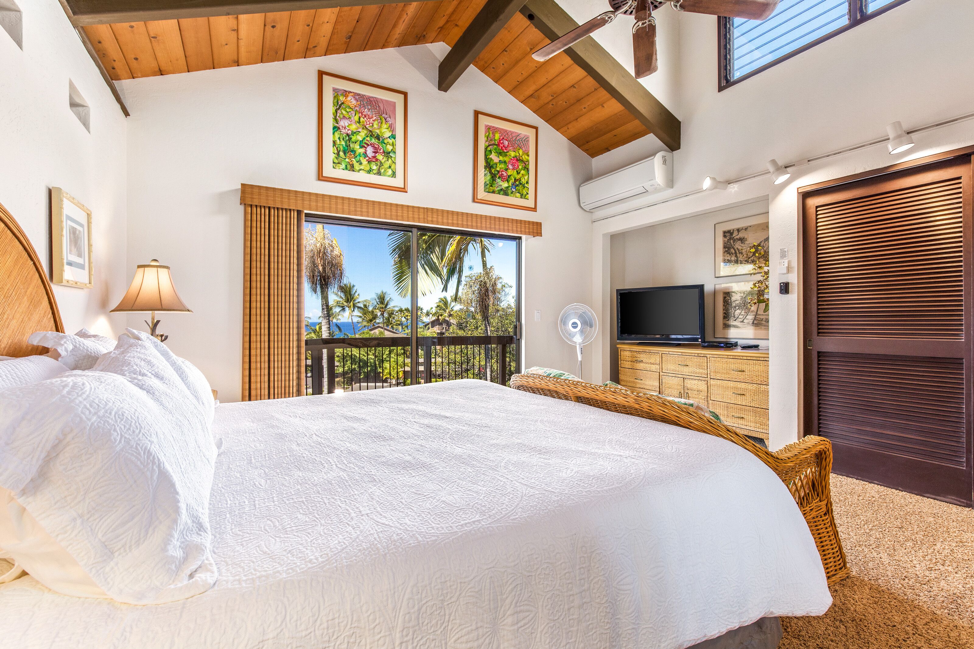 A bedroom with a large bed, a TV, a balcony with a view of palm trees, and a wooden ceiling.
