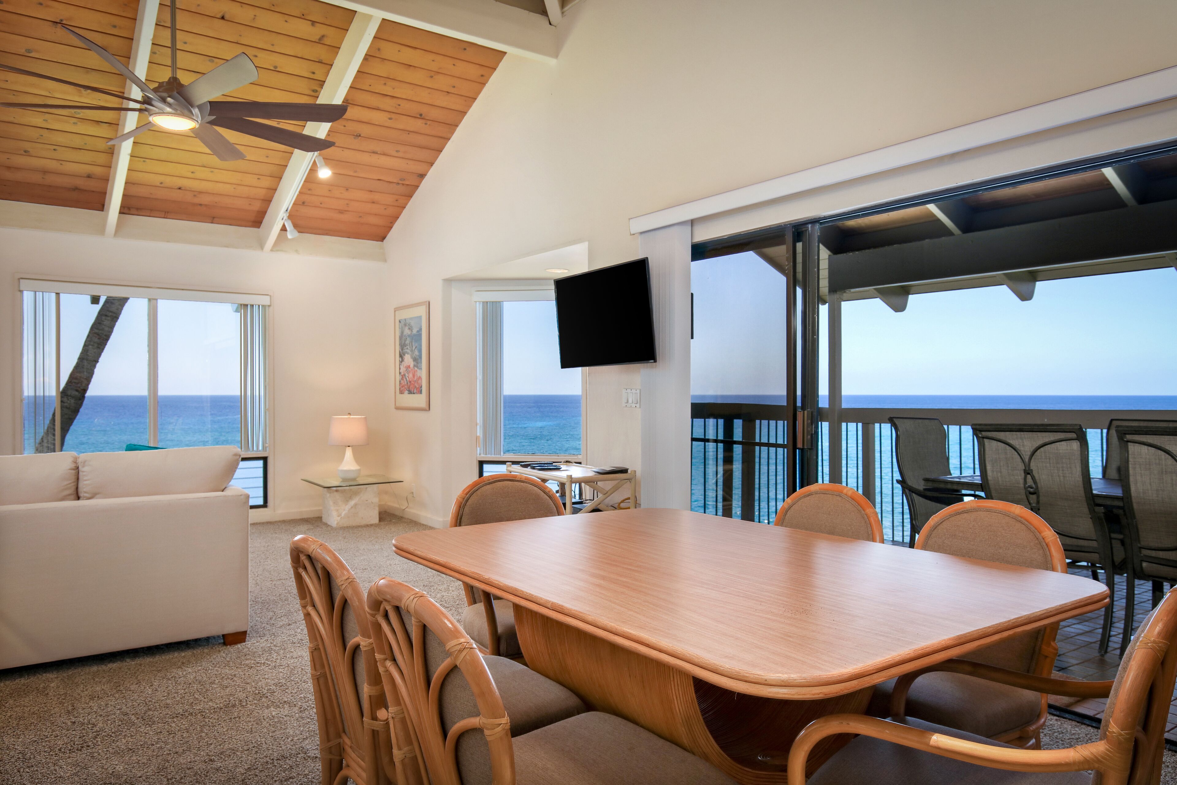 A spacious living area with a wooden dining table, comfortable seating, a ceiling fan, a flat-screen TV, and a large window overlooking the ocean.