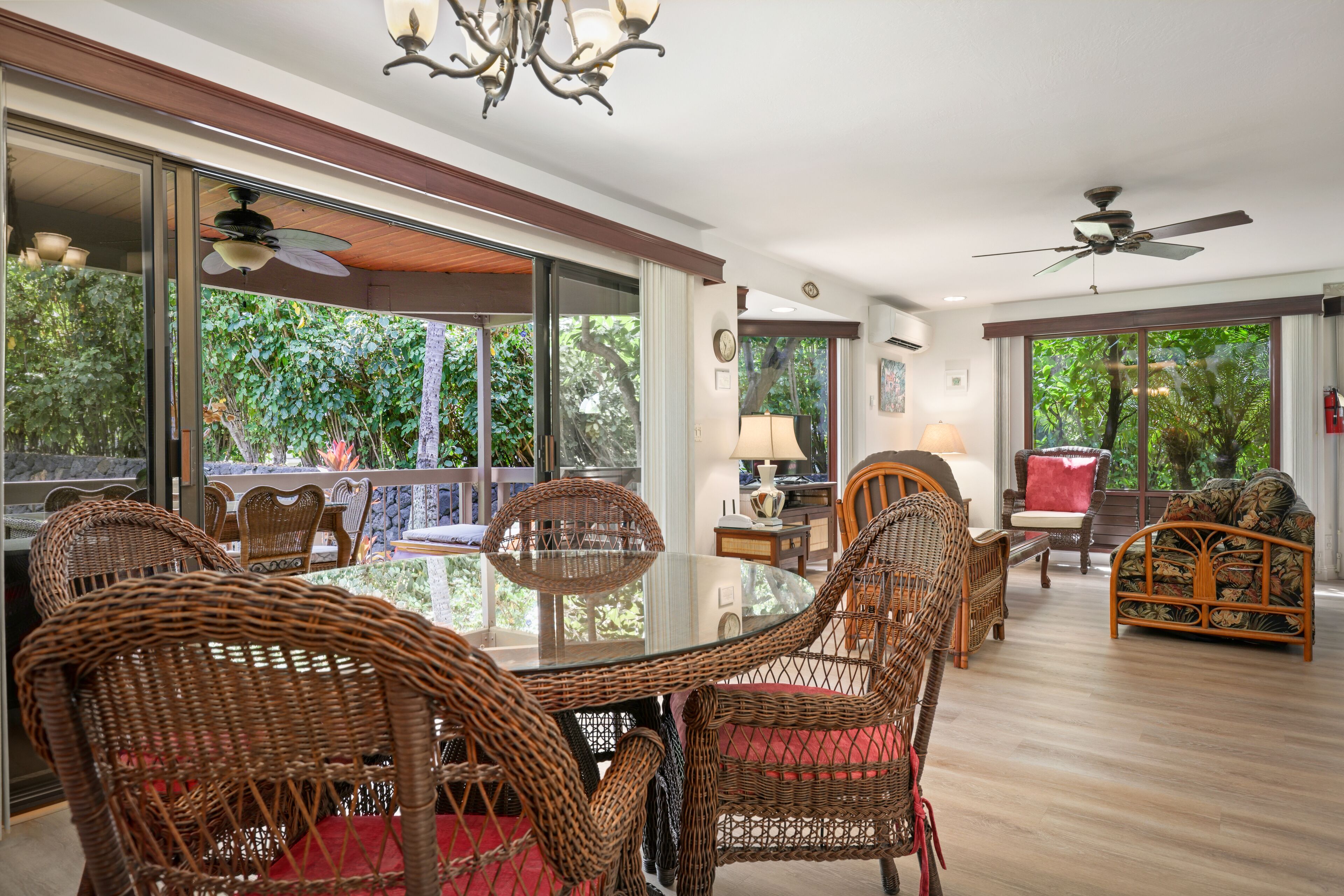 A spacious room with a dining table surrounded by wicker chairs, a ceiling fan, and large windows offering a view of greenery outside.