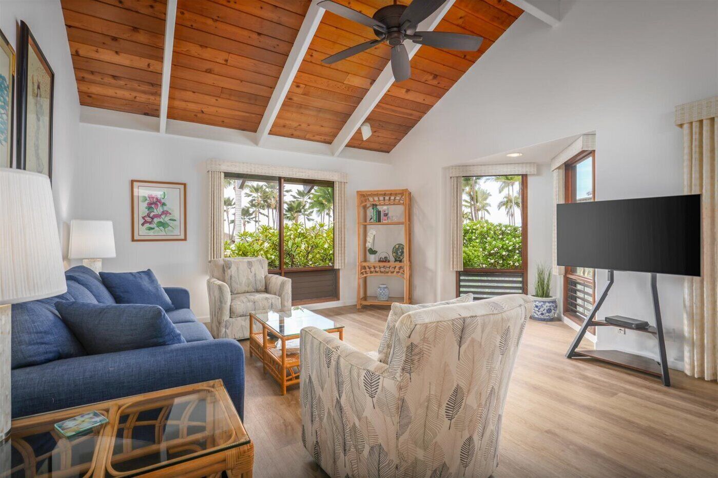 A spacious living room with a high ceiling, wooden beams, a ceiling fan, a blue sofa, patterned armchairs, a coffee table, a TV on a stand, and large windows with a view of greenery.