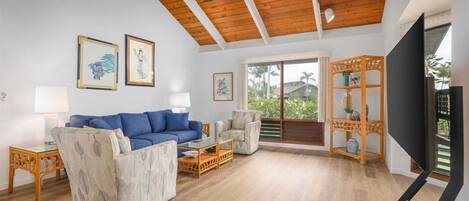 A spacious living room with a blue sofa, a patterned armchair, a glass coffee table, and a TV on a stand.