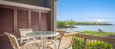 A covered outdoor area with a round table and chairs, overlooking a coastal view with buildings and a clear sky.
