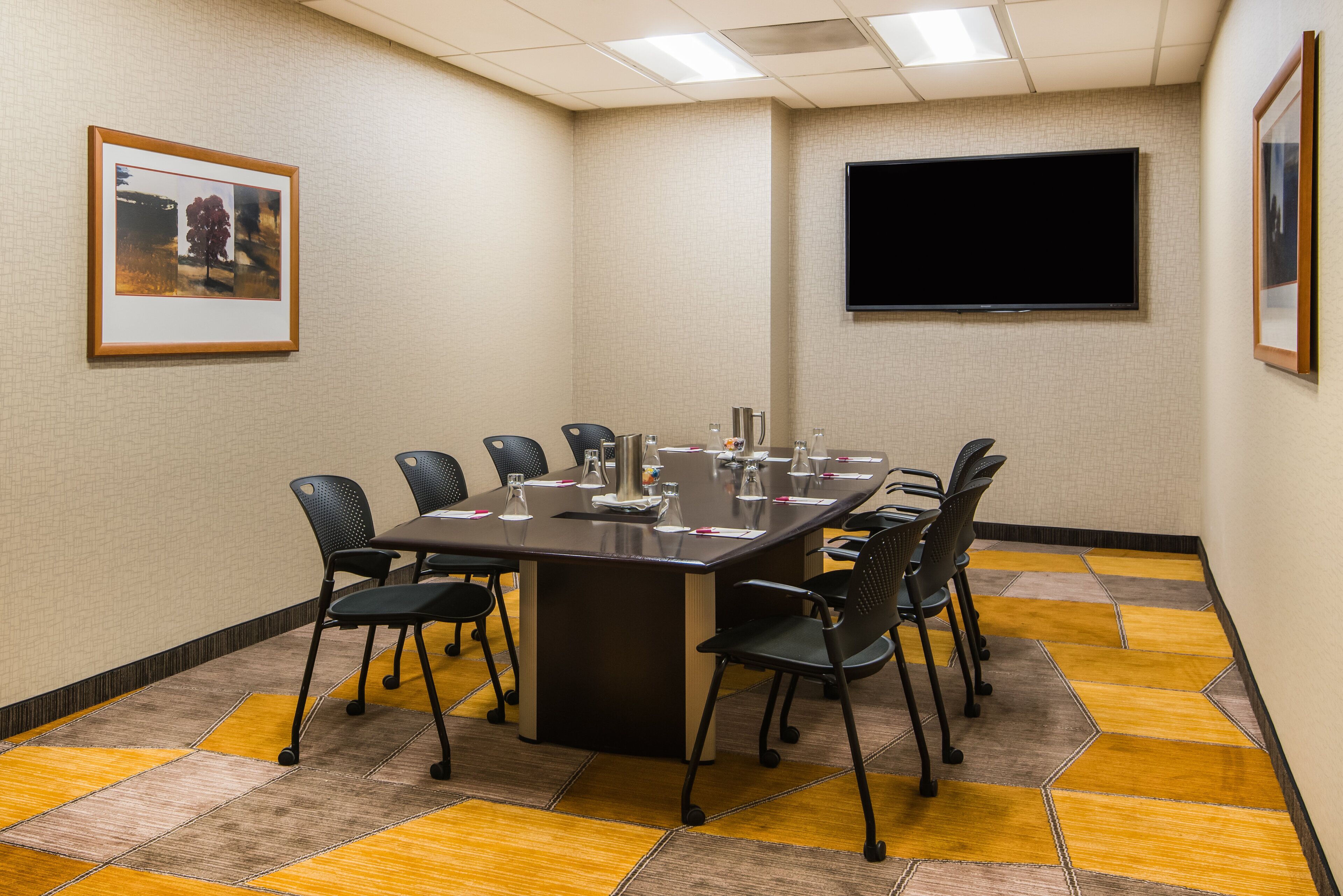 A conference room with a long table, six chairs, a flat-screen TV, and framed artwork on the walls.