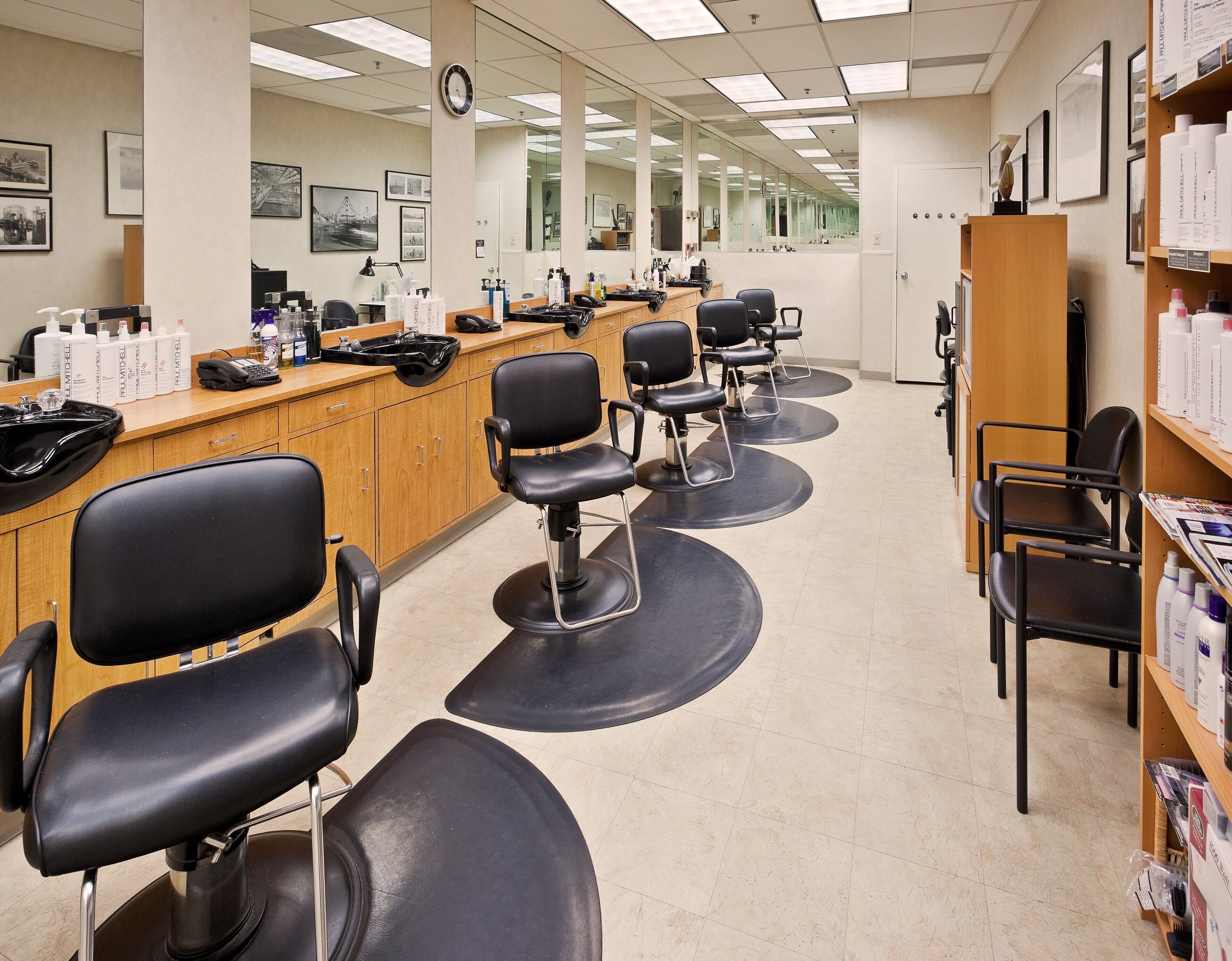 A salon with multiple black salon chairs, a wooden counter with various hair products, and framed pictures on the walls.