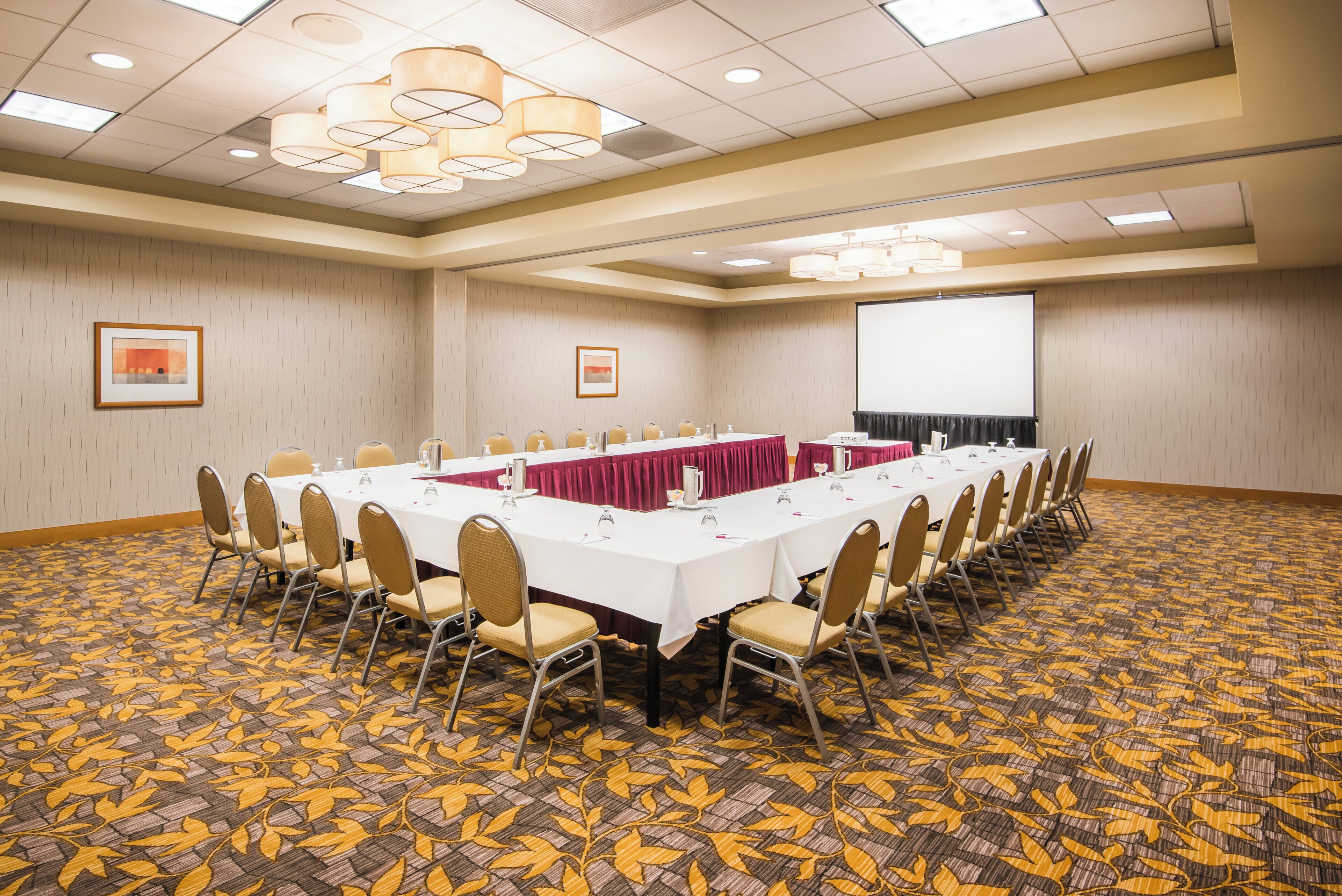 A conference room with a long table, chairs, a projector screen, and a patterned carpet.