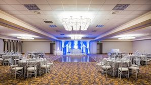 Une grande salle de banquet avec des tables rondes et des chaises, une scène avec des rideaux bleus et un tapis à motifs.