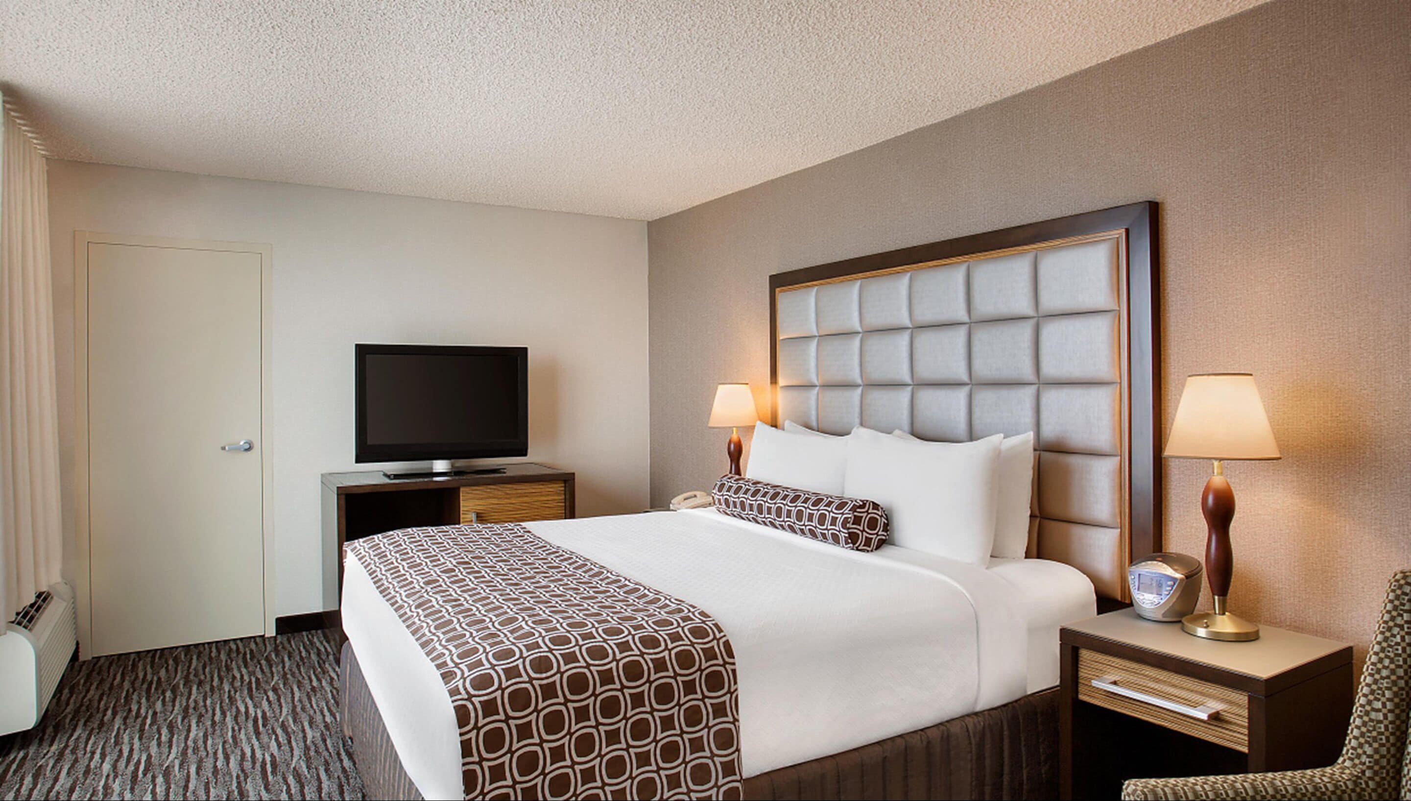A hotel room with a large bed, a television on a stand, two bedside lamps, a desk with a lamp, and a chair.