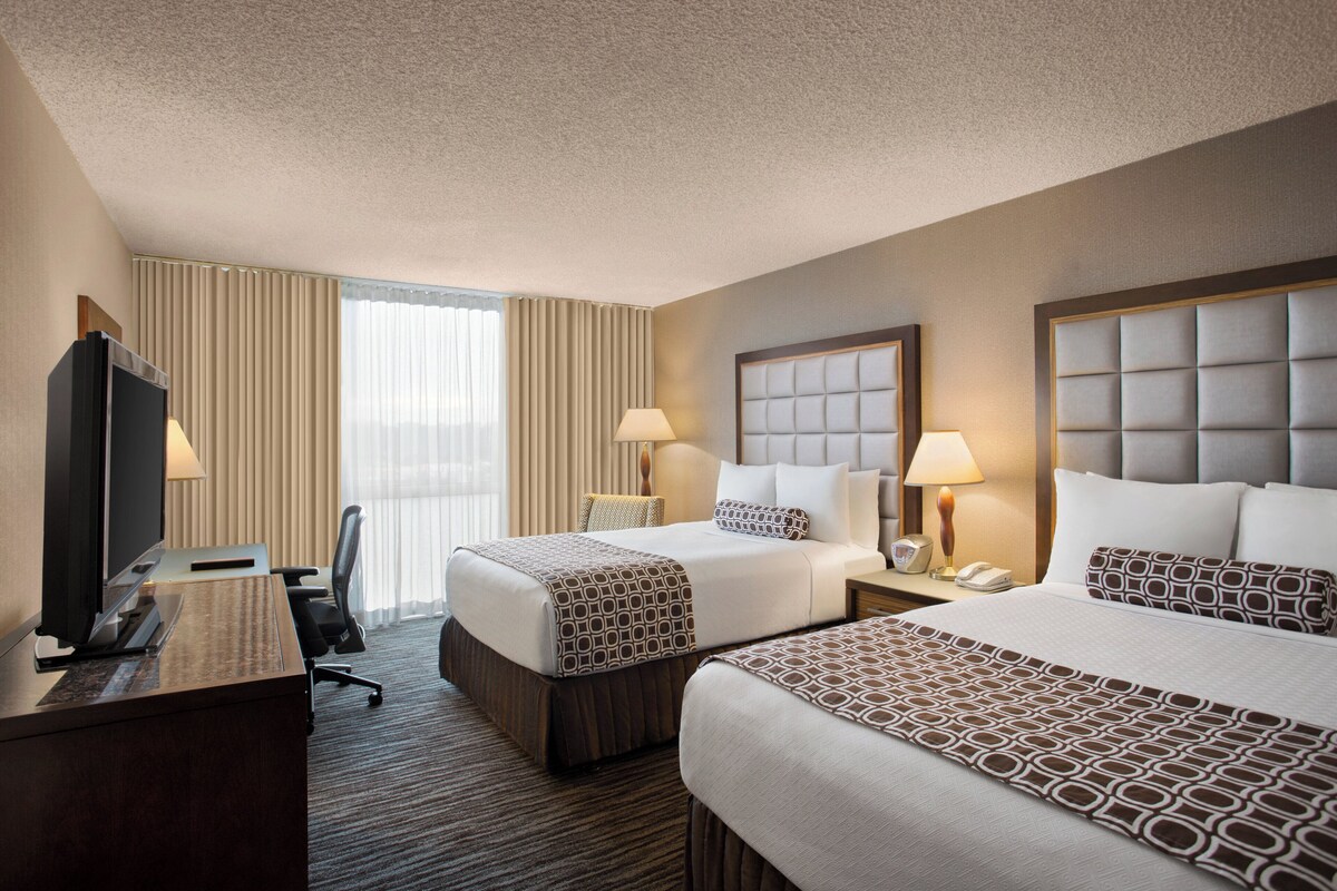 A hotel room with two beds, a desk, a TV, and a window with curtains.