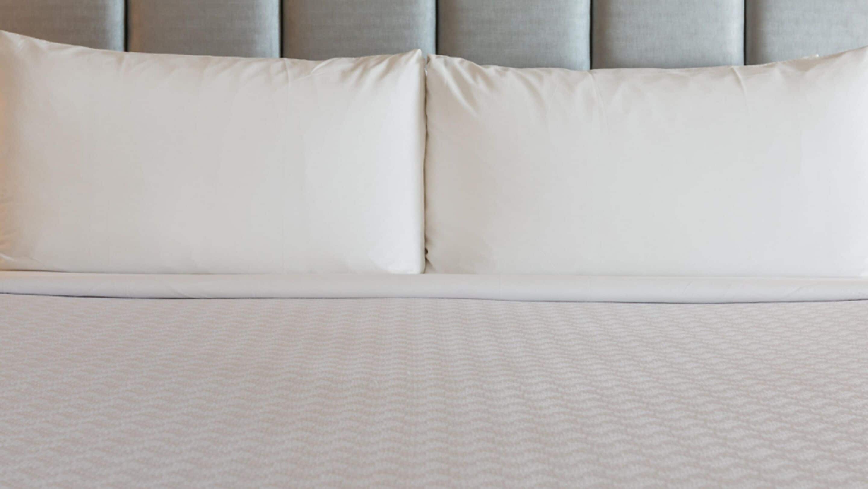 A neatly made bed with a white duvet and pillows, a grey upholstered headboard, and a textured white bedspread.