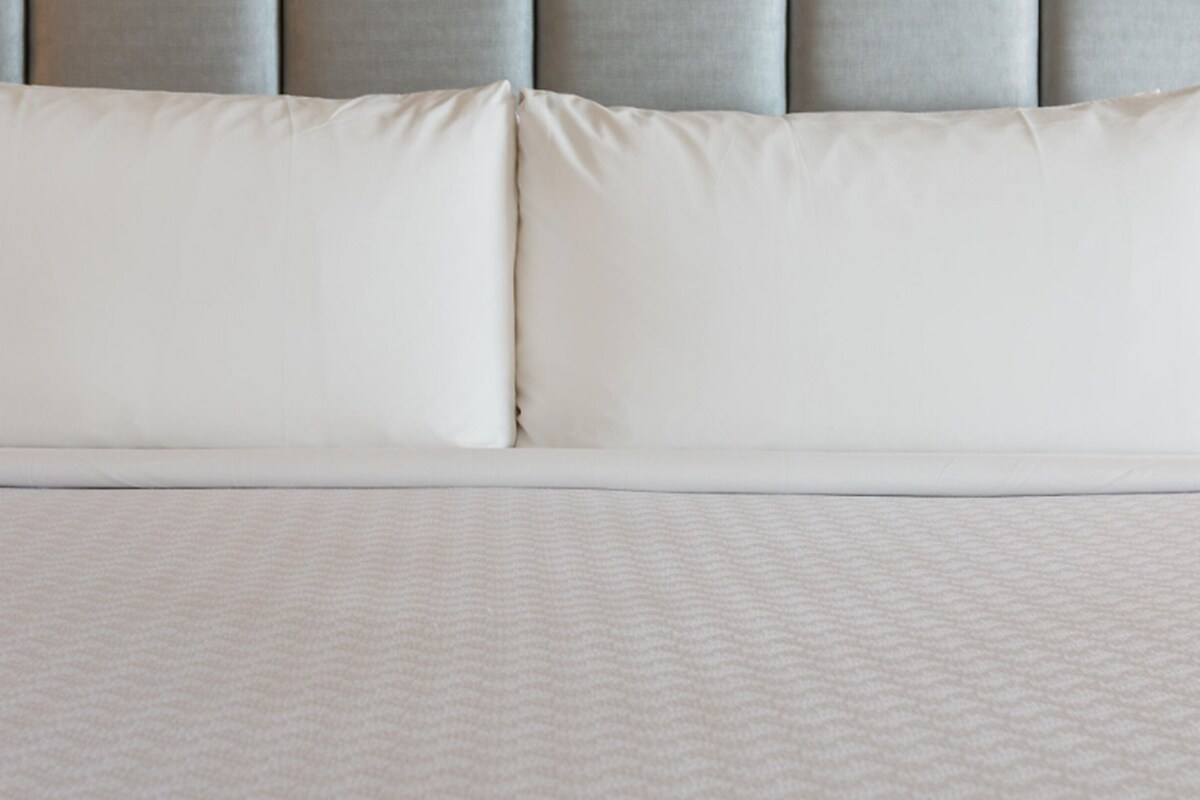 A neatly made bed with a white duvet and pillows, a grey upholstered headboard, and a textured white bedspread.