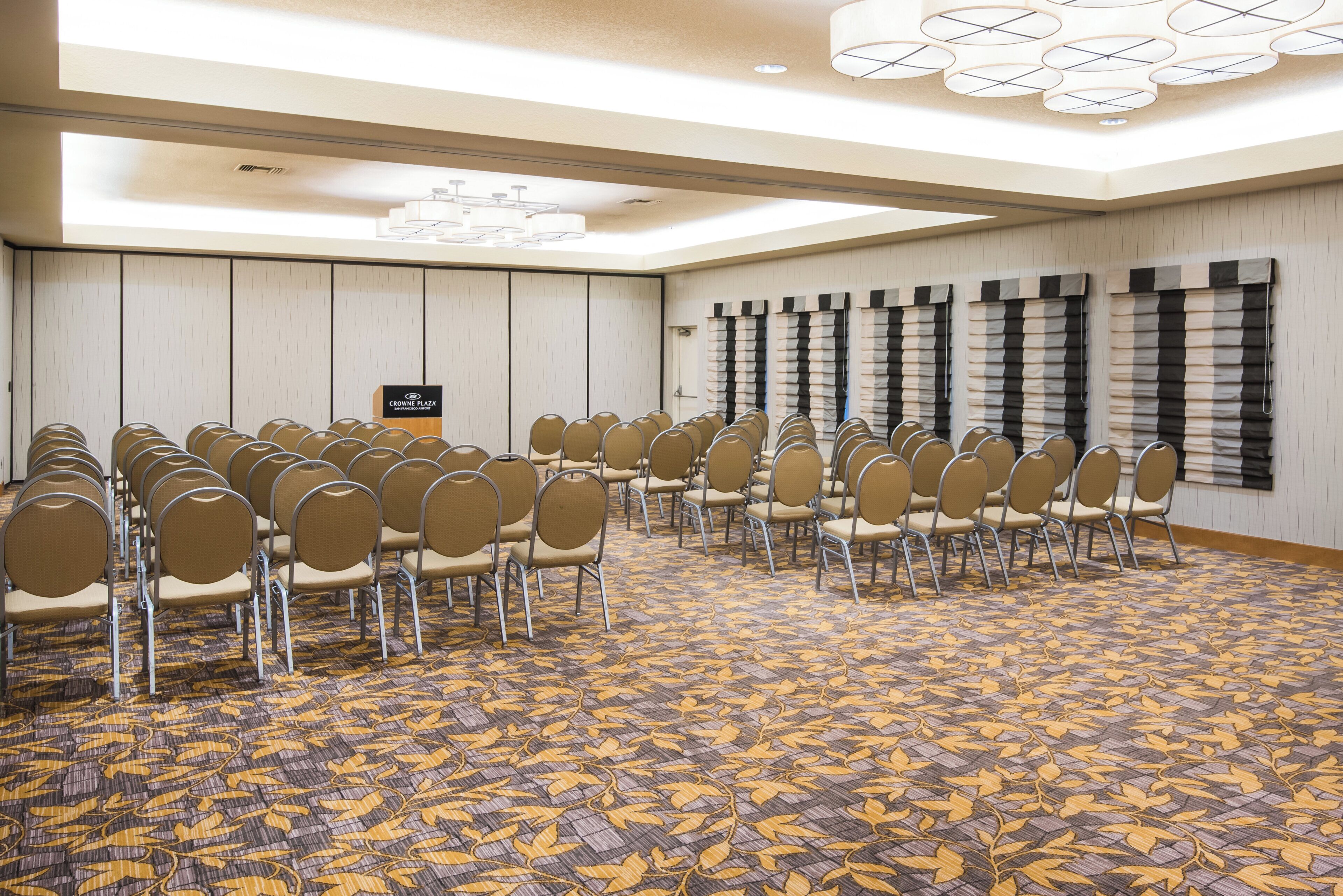 Un salón de conferencias con hileras de sillas, una alfombra estampada y un cartel que indica que forma parte del Crowne Plaza San Francisco Airport.
